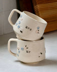 Two ceramic mugs, one labeled 'Mrs.' and the other 'Mr.', stacked on a light surface with a wooden board in the background.