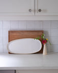 White oval platter on a wooden cutting board with a small vase of flowers on a tiled kitchen counter.