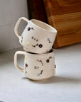 Two stacked ceramic mugs with floral designs and text on a light surface.