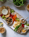 Platter of assorted cheeses, crackers, and vegetables on a white surface with a cutting board and additional food items.