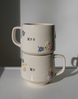 Two stacked ceramic mugs labeled 'Mrs' and 'Mr' with floral designs on a plain background.