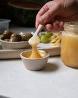 Hand using a small spoon to scoop a yellow condiment into a ceramic dish on a table with olives and bread.