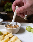 Hand holding a small spoon over a bowl of spreadable food with cheese and grapes on a plate.