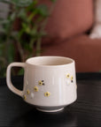 A cream-colored ceramic mug with small yellow and white floral details, sitting on a dark wooden table with a blurred background of a sofa and green plant.