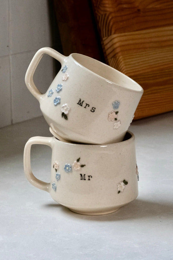 Two ceramic mugs, one labeled 'Mrs.' and the other 'Mr.', stacked on a light surface with a wooden board in the background.