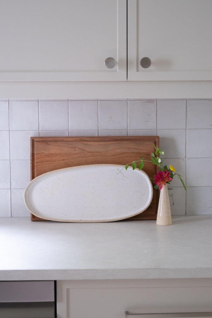 White oval platter on a wooden cutting board with a small vase of flowers on a tiled kitchen counter.