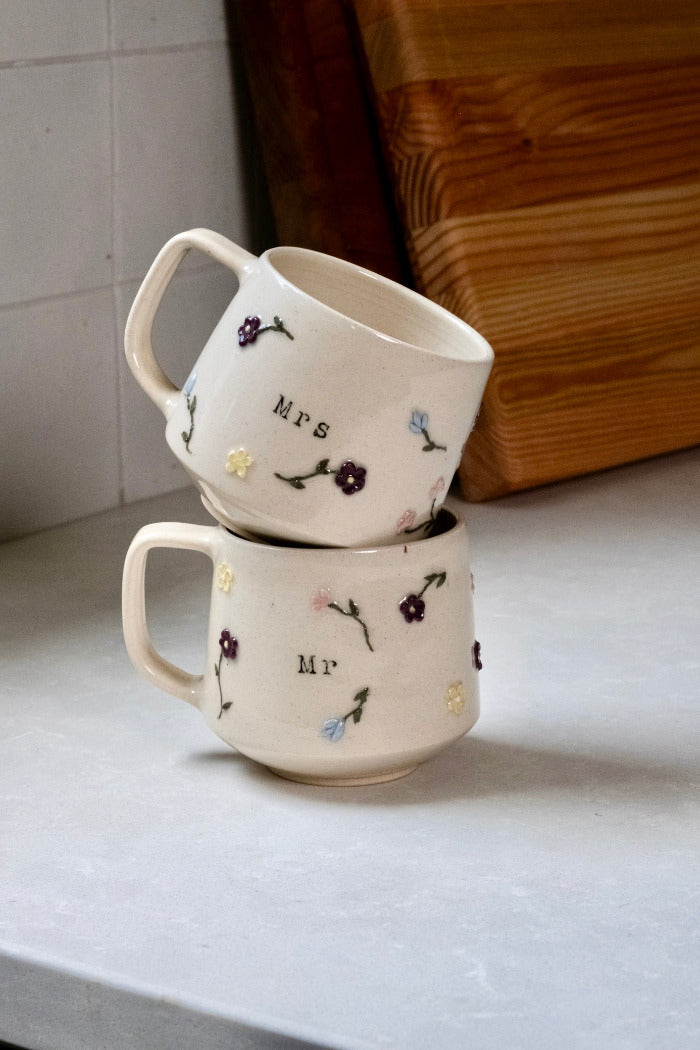 Two stacked ceramic mugs with floral designs and text on a light surface.