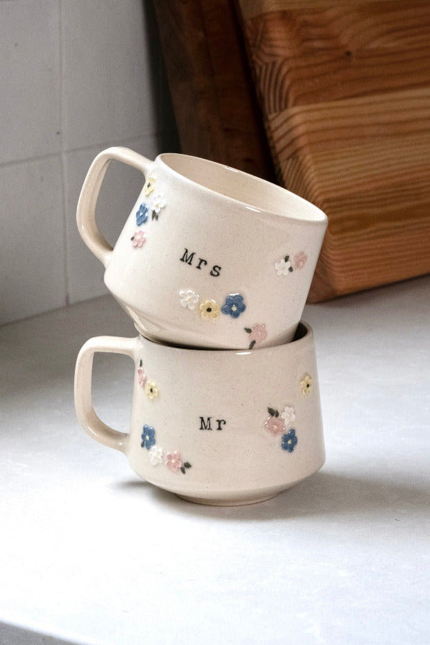 Two stacked ceramic mugs with 'Mrs.' and 'Mr.' text on a white surface.