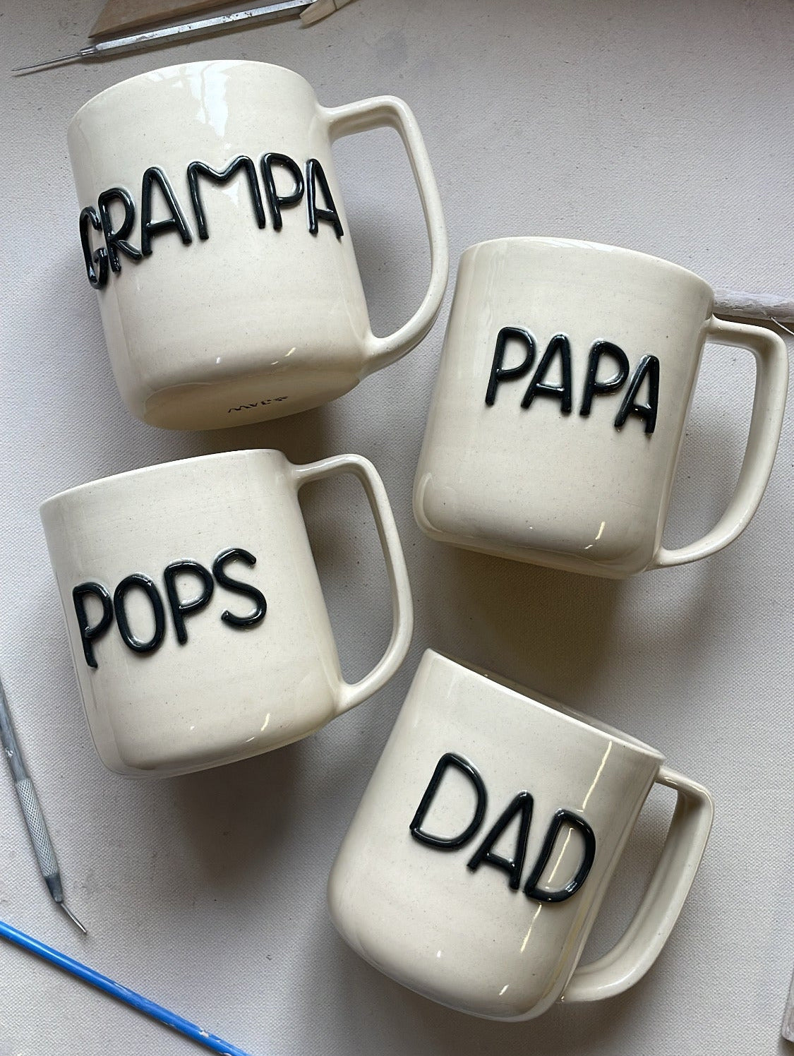 Four white mugs with 'GRAMPA', 'PAPA', 'POPS', and 'DAD' text on a light gray background.