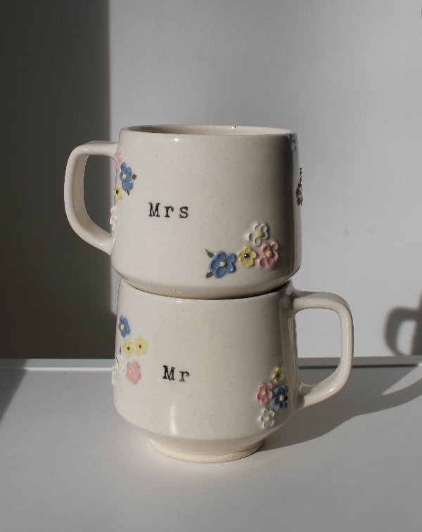 Two stacked ceramic mugs labeled 'Mrs' and 'Mr' with floral designs on a plain background.