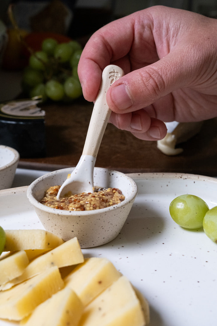 Hand holding a small spoon over a bowl of spreadable food with cheese and grapes on a plate.