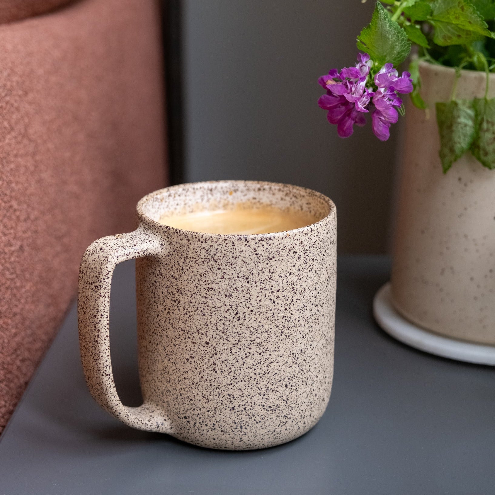 Speckled ceramic mug with a hot beverage on a surface next to a potted plant.