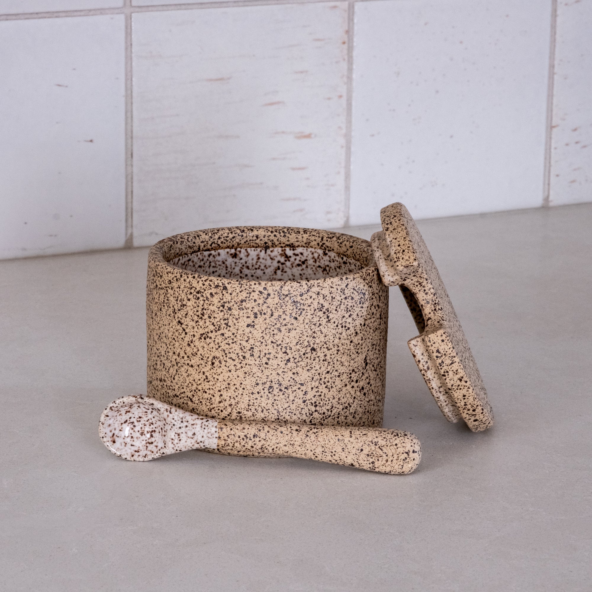 Speckled ceramic jar with lid and scrub brush on a light surface with tiled wall background