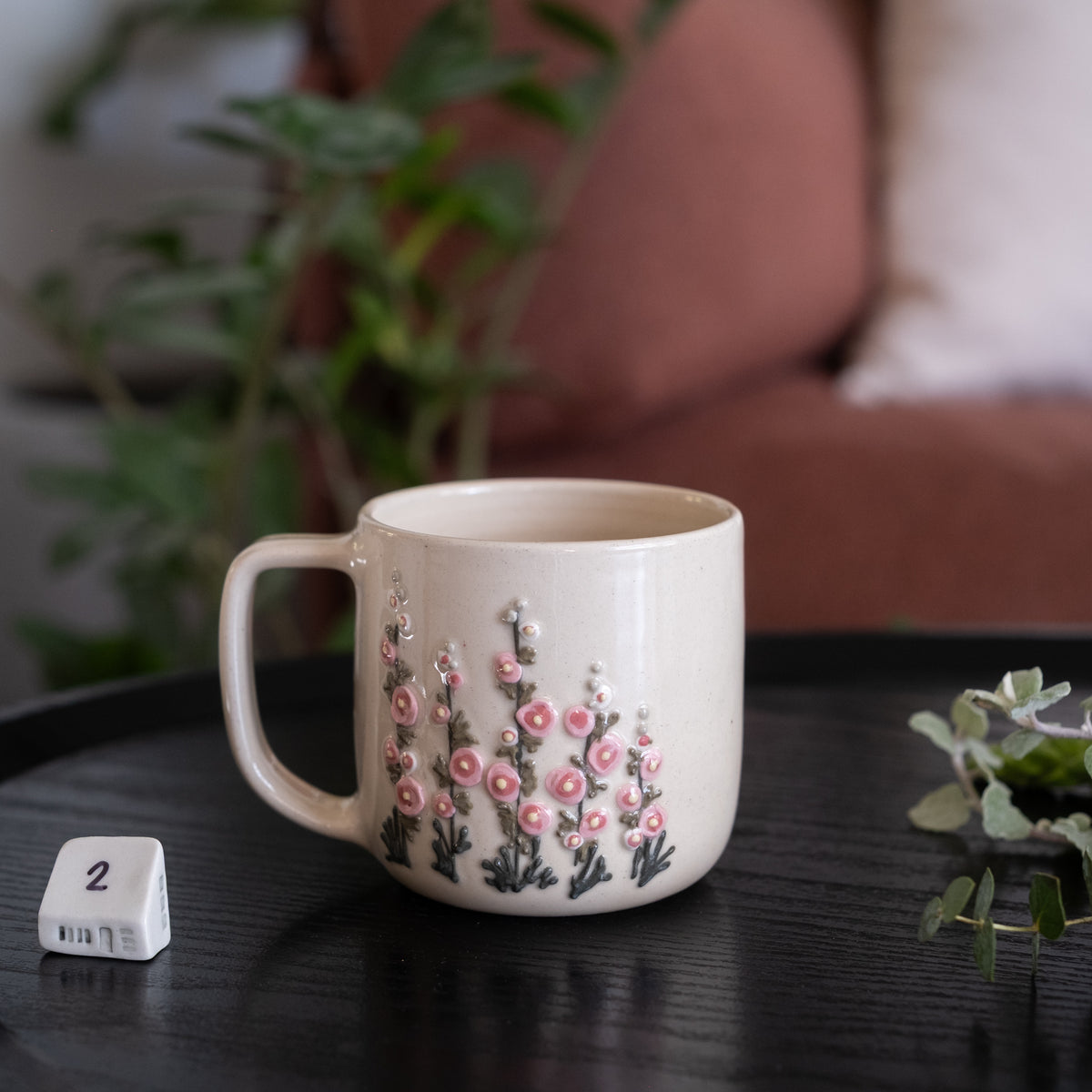 A cream-colored ceramic mug with a pink and green floral pattern, sitting on a dark wooden table next to a white dice cube in a cozy, softly blurred indoor setting.