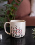 A cream-colored ceramic mug with a pink and green floral pattern, sitting on a dark wooden table next to a white dice cube in a cozy, softly blurred indoor setting.