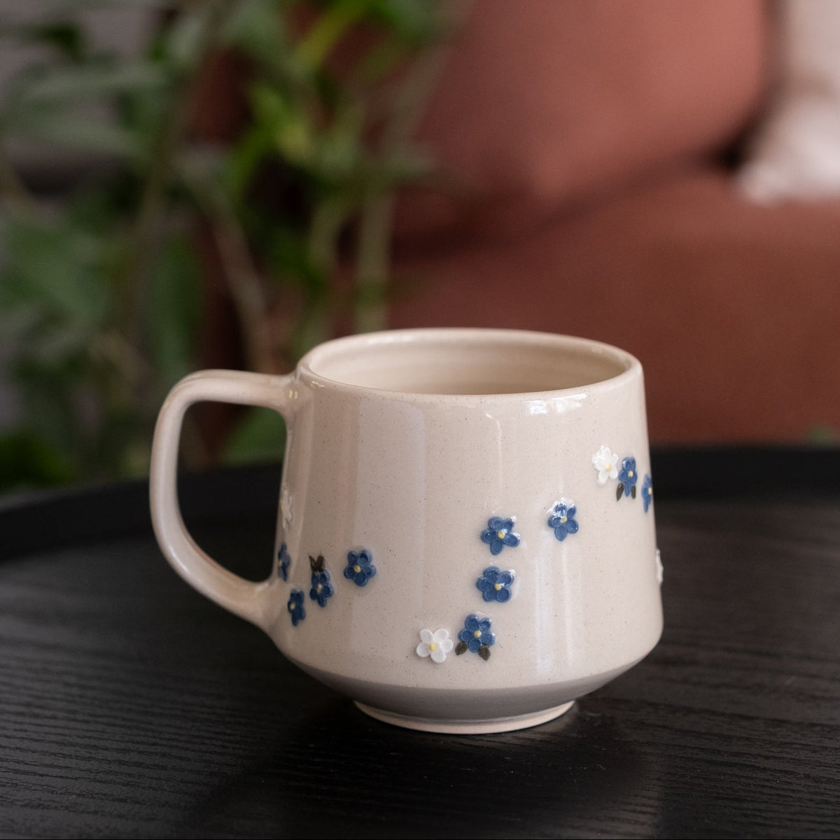 A cream-colored ceramic mug with small blue and white floral patterns, resting on a dark wooden table with a blurred background of a sofa and green plant.