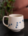 A cream-colored ceramic mug with small blue and white floral patterns, resting on a dark wooden table with a blurred background of a sofa and green plant.