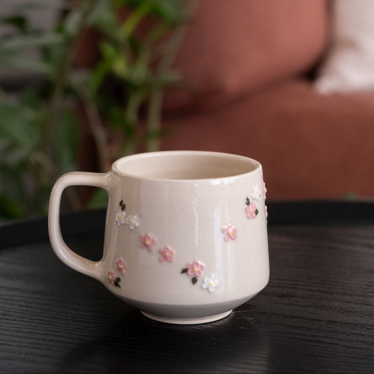 A cream-colored ceramic mug with a handle, decorated with small pink and white floral details, resting on a dark wooden table with a blurred background of a plant and a sofa.