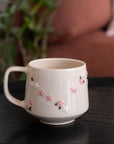 A cream-colored ceramic mug with a handle, decorated with small pink and white floral details, resting on a dark wooden table with a blurred background of a plant and a sofa.